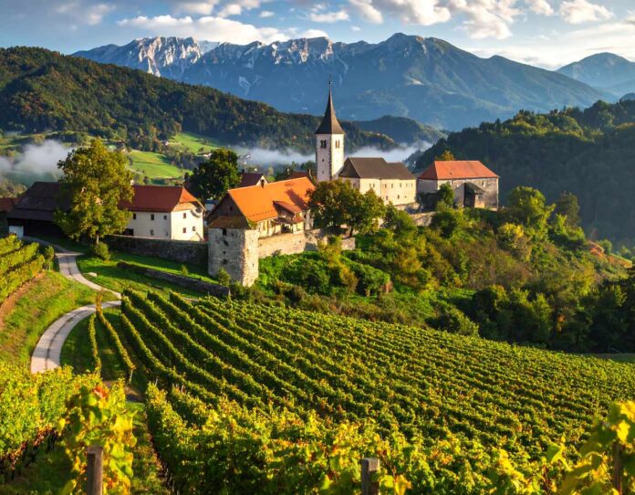 A scenic village nestles among verdant vineyards, with a church steeple rising against majestic mountains and a partly cloudy sky